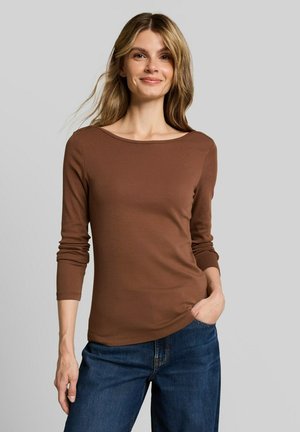 Vrouw met lichtbruin haar, gekleed in een bruin shirt met lange mouwen en een blauwe spijkerbroek, staat met één hand in haar zak en glimlacht.