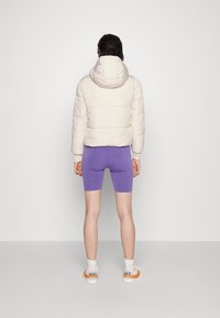 Kvinde står med ryggen til kameraet iført en hvid polstret jakke, lilla cykelshorts, hvide sokker og hvide Nike sneakers med orange swoosh.