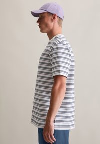 Gestreiftes Kurzarm-T-Shirt in Weiß und Marineblau mit Rundhalsausschnitt, getragen mit einer lila Mütze. Die Seitenansicht zeigt den lässigen Schnitt und die Stofftextur.