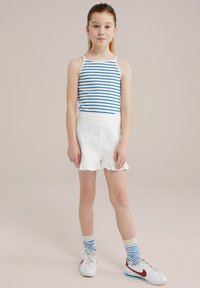 Pasi rebrasto belo in modro majico brez rokavov, v kombinaciji z belimi šortsi s čipkastim robom. Bele superge z rdečimi poudarki in modrimi rebrastimi nogavicami.