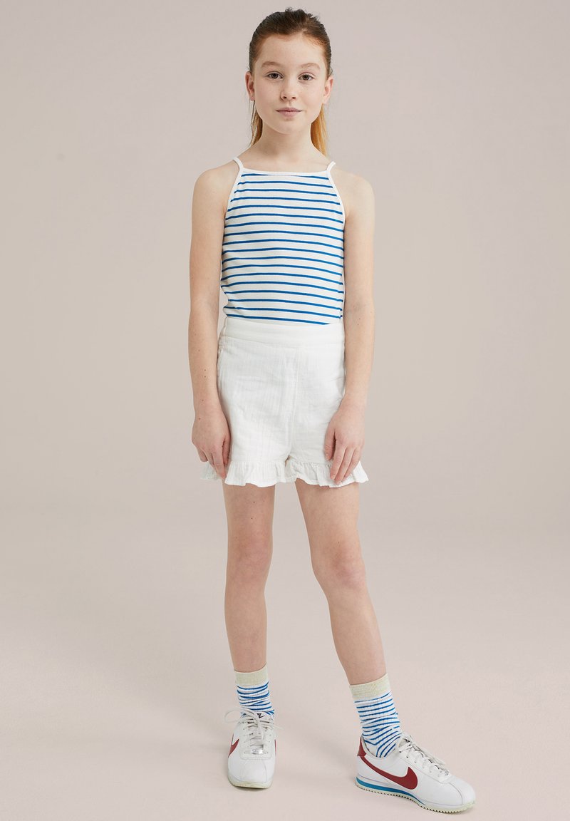 Pasi rebrasto belo in modro majico brez rokavov, v kombinaciji z belimi šortsi s čipkastim robom. Bele superge z rdečimi poudarki in modrimi rebrastimi nogavicami.