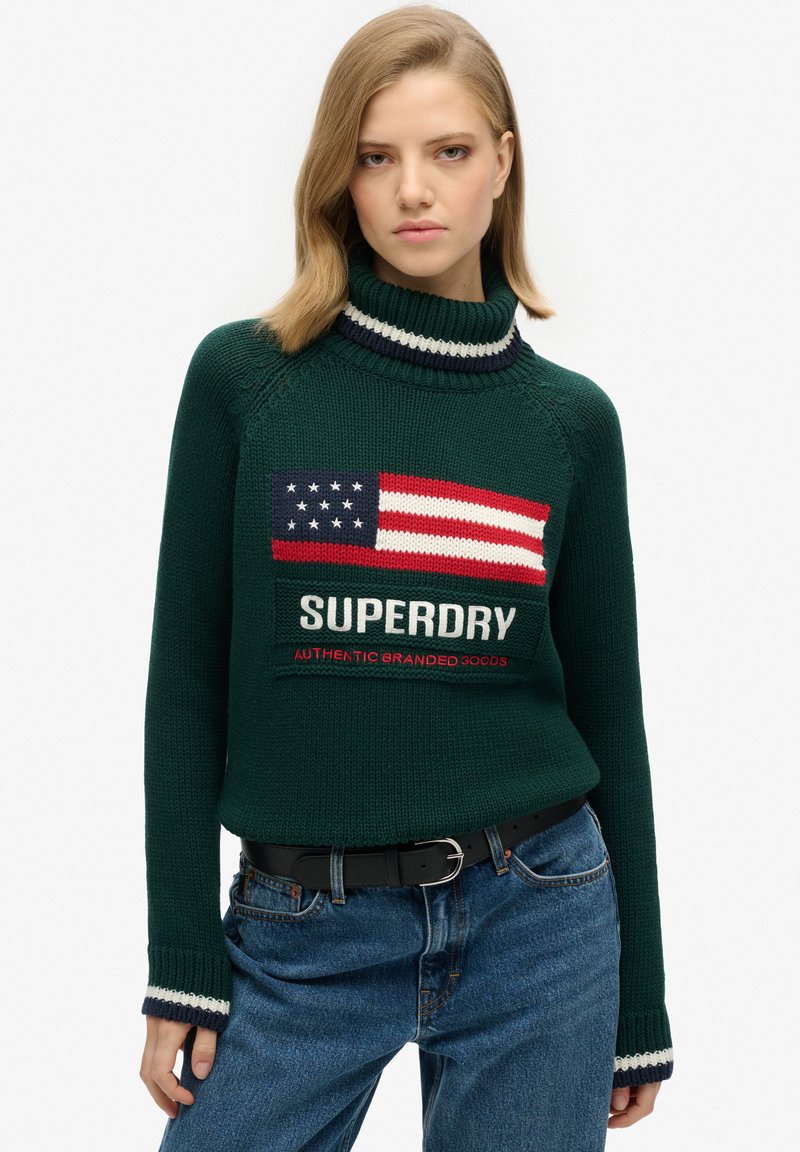 Pull en maille vert avec col roulé, présentant un motif de drapeau américain et "SUPERDRY" en texte blanc. Accentué par des rayures bleu marine et blanches.