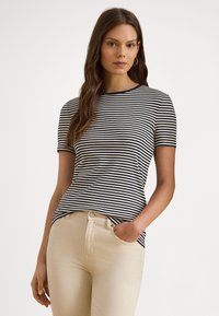 Sieviete tērpusies tumši zilā un baltā svītrainā īsroku krekliņā un beige krāsas biksēs, stāvot ar vienu roku kabatā uz vienkārša fona.