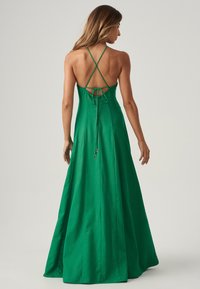 Abito maxi verde con corpetto aderente, sottili spalline incrociate e un laccetto regolabile sul retro; realizzato in tessuto morbido e fluido.