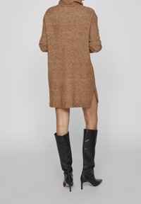Abito a maglia color marrone caldo con collo alto, spacchi laterali e vestibilità rilassata. Abbinato a stivali neri in pelle con tacco a spillo fino al ginocchio.
