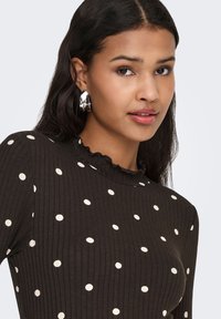 Mørkebrun ribbet top med hvide polkaprikker, med en flæset halsudskæring og lange ærmer. Sølv øreringe i hoop-stil bæres.