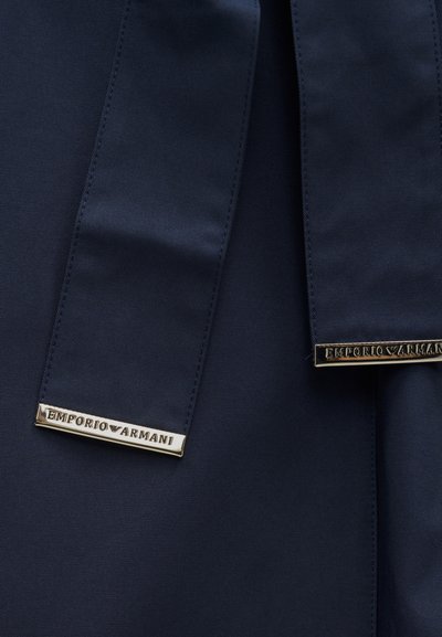 Υφασμα σε ναυτικό μπλε με ομαλή υφή, διακοσμημένο με δύο χρυσές λεπτομέρειες με την επιγραφή "EMPORIO ARMANI", τοποθετημένες σε ραμμένη ραφή, τονίζοντας την επωνυμία.