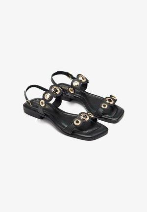 Sandalias de cuero negro con punta cuadrada y un tacón bajo, que presentan acentos circulares en tono dorado y correas ajustables en el tobillo.