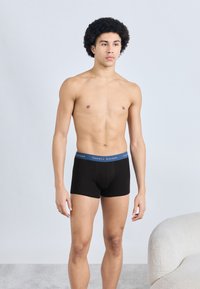 Μαύρα ανδρικά μποξεράκια με μπλε μέση που φέρει το λογότυπο "Tommy Hilfiger", παρουσιασμένα σε ανδρικό μοντέλο μπροστά σε ουδέτερο φόντο.