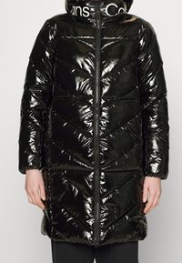 Svart puffjacka med en glansig, kviltad yta, med dragkedja fram, huva och chevronmönster. Kragen har vita varumärkesdetaljer.