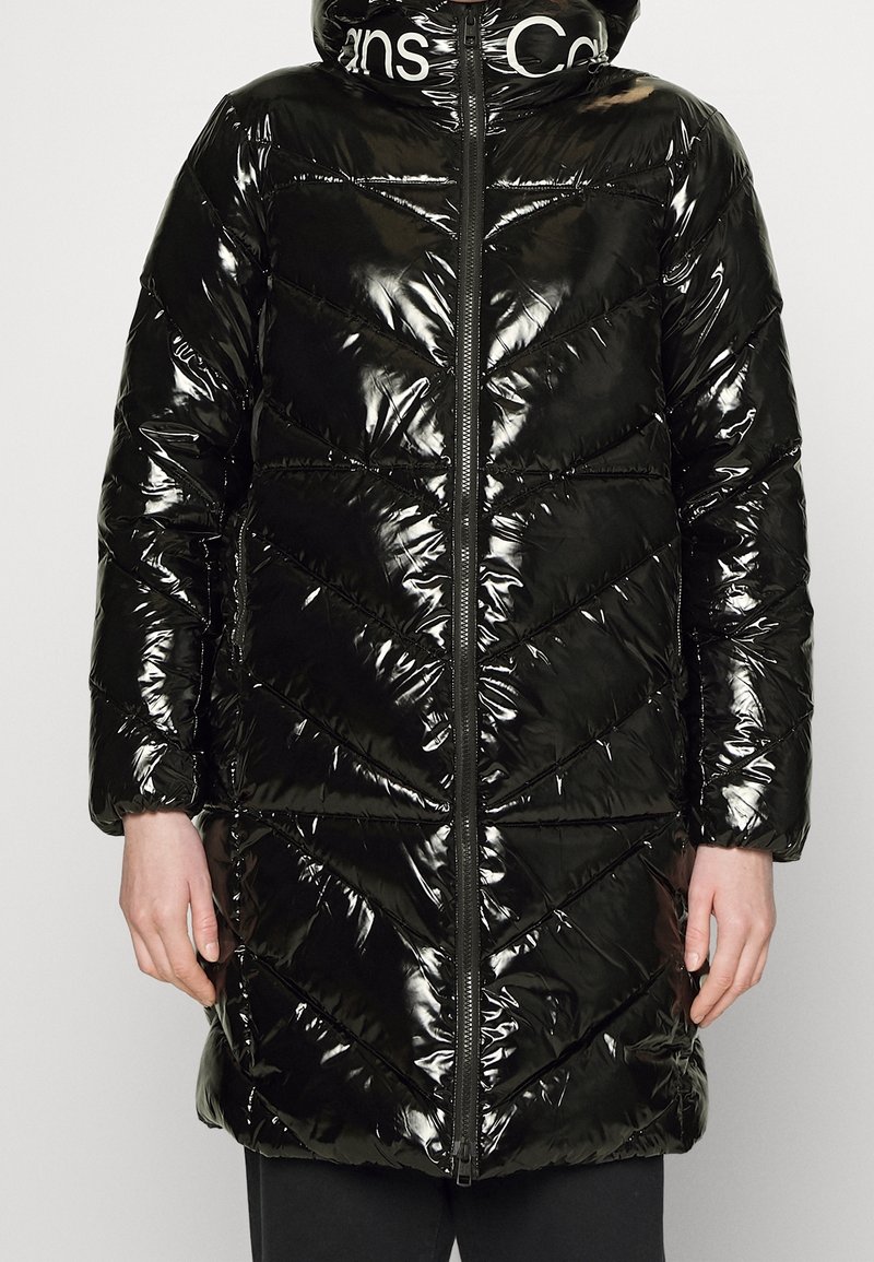 Svart puffjacka med en glansig, kviltad yta, med dragkedja fram, huva och chevronmönster. Kragen har vita varumärkesdetaljer.