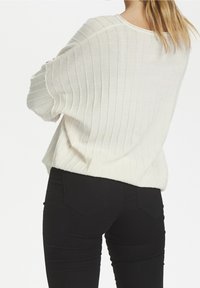 Weißer gerippter Strickpullover mit lockerer Passform und weiten Ärmeln, kombiniert mit engen schwarzen Jeans, die ein einfaches Design mit Gesäßtaschen aufweisen.