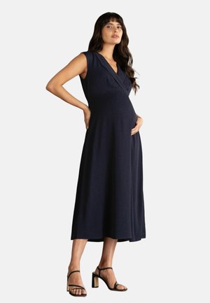 Zwangere vrouw draagt een mouwloze, marineblauwe midi-jurk met een V-hals en zwarte sandalen met blokhak en bandjes, staand met één hand op de buik.