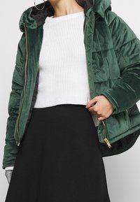 Kvinna i en mörkgrön sammetsjacka med vaddering över en vit stickad tröja och en svart kjol, med gyllene örhängen som syns.