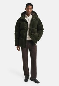 Dunkelgrüne Cordura-Steppjacke mit Kapuze, Druckknöpfen und zwei Taschen vorne, kombiniert mit einem hellen Hemd und braunen Hosen.