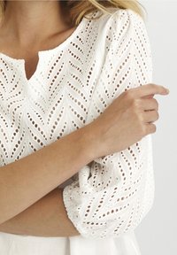 Donna che indossa una camicia bianca con un intricato motivo in pizzo a occhiello, maniche raccolte ai polsi e un piccolo scollo a V.