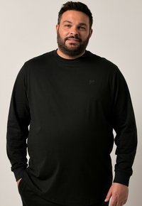 Hombre con pelo corto y oscuro y barba, vistiendo una camisa de manga larga negra, de pie con las manos en los bolsillos contra un fondo claro y liso.