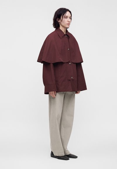 Personne portant une veste cape ample bordeaux foncé sur un pantalon large beige clair et des chaussures noires, debout devant un fond blanc uni.