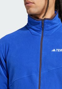 Blue fleece jacket with high collar and zip closure. Features a contrasting dark zipper and logo on the chest. Soft, textured fabric.