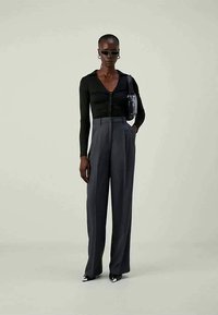 Pessoa alta vestindo uma blusa preta de manga longa ajustada, calças largas cinzas de cintura alta, sapatos pontiagudos pretos e brancos, óculos de sol e uma bolsa de ombro.