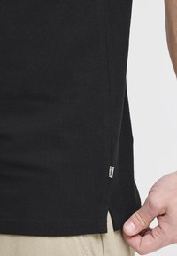 Sort bomulds T-shirt med struktureret væv, der har sideskår og et lille mærke nær kanten. Skåret til en afslappet pasform.