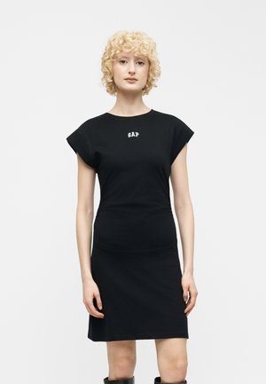 Femeie cu păr scurt și creț, blond, purtând o rochie neagră cu mâneci scurte, cu logo-ul „GAP” pe piept, stând în fața unui fundal alb simplu.