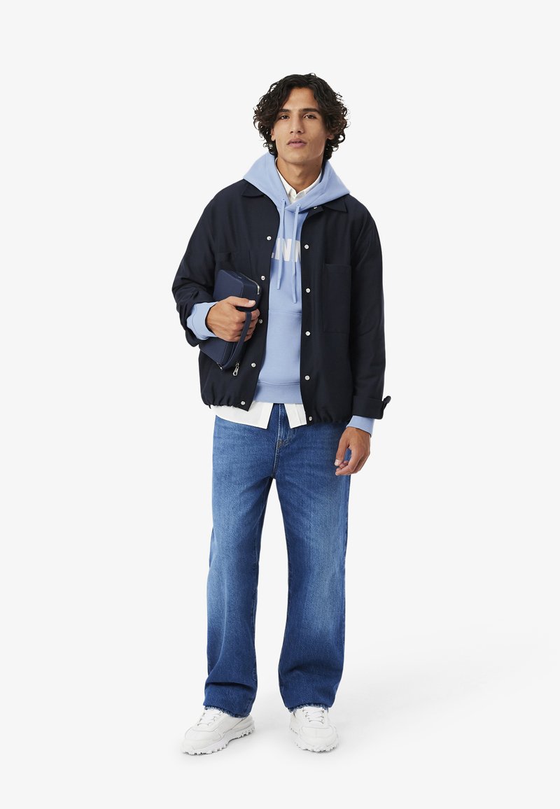Uomo con capelli ricci che indossa una giacca nera sopra una felpa azzurra chiara, camicia bianca, jeans blu, sneakers bianche, che tiene in mano una pochette blu scuro.