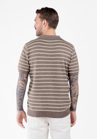 Hombre con brazos tatuados lleva una camisa de manga corta a rayas marrón y beige y pantalones blancos, de espaldas contra un fondo liso.