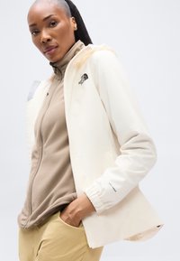 Αδιάβροχο μπουφάν κρεμ χρώματος με κουκούλα, διαθέτει ελαστικές μανσέτες και φερμουάρ. Φορεμένο πάνω από μπεζ φλις τοπ, συνδυασμένο με παντελόνι σε ταμπά απόχρωση.