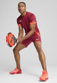 Raudonas ir oranžinis sportinis marškinėliai ir šortai su lygiu paviršiumi, laiko juodai raudoną raketę, vilkėdamas oranžinius sportinius batus.