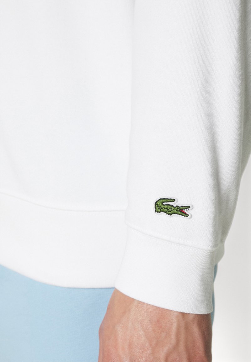Sweatshirt blanc avec des poignets côtelés, comportant un petit logo de crocodile brodé en vert sur la manche gauche. Matière en coton lisse.