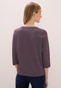 Kvinna med ljusbrunt hår som bär en lila och vit randig topp med 3/4 ärmar och mörkblå jeans, stående mot en neutral bakgrund.
