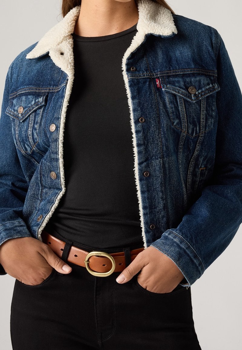 Personne portant une veste en jean bleu foncé avec doublure sherpa blanche, chemise noire à côtes, pantalon noir et ceinture en cuir marron avec boucle ronde dorée.