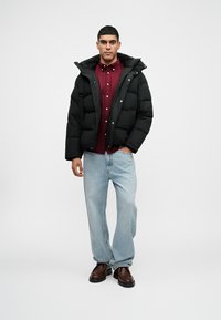 Jachetă puffer neagră cu glugă, având panouri mari matlasate. Sub ea, o cămașă burgundy cu nasturi și blugi bleu deschis, lejeri. Pantofi maro.