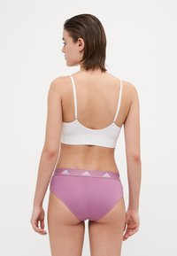 Bílý žebrovaný bralet s nastavitelnými ramínky, doplněný fialovými kalhotkami s vysokým pasem a logovaným pasem. Hladká textura.
