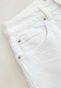 Gros plan sur un jean en denim blanc montrant la poche avant, la passants de ceinture et les rivets métalliques sur un fond clair.