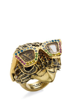 Goudkleurige ring in de vorm van een ingewikkeld gezicht met kleurrijke brillenglazen in kattenoogstijl bezaaid met edelstenen, met de tekst "Kurt Geiger London" gegraveerd aan de binnenkant.