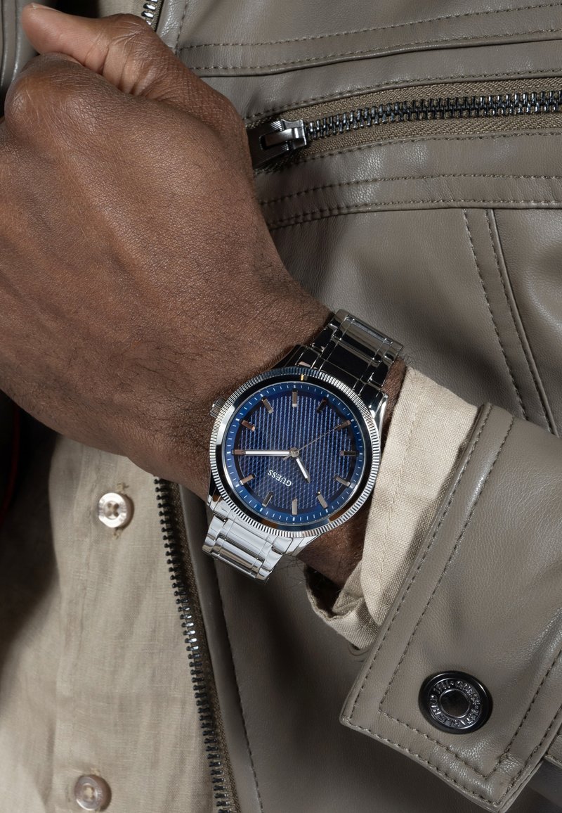 Montre en argent avec un cadran texturé bleu, aiguilles et index argentés, bracelet métallique, portée sur une main avec une veste en cuir et une chemise visibles.