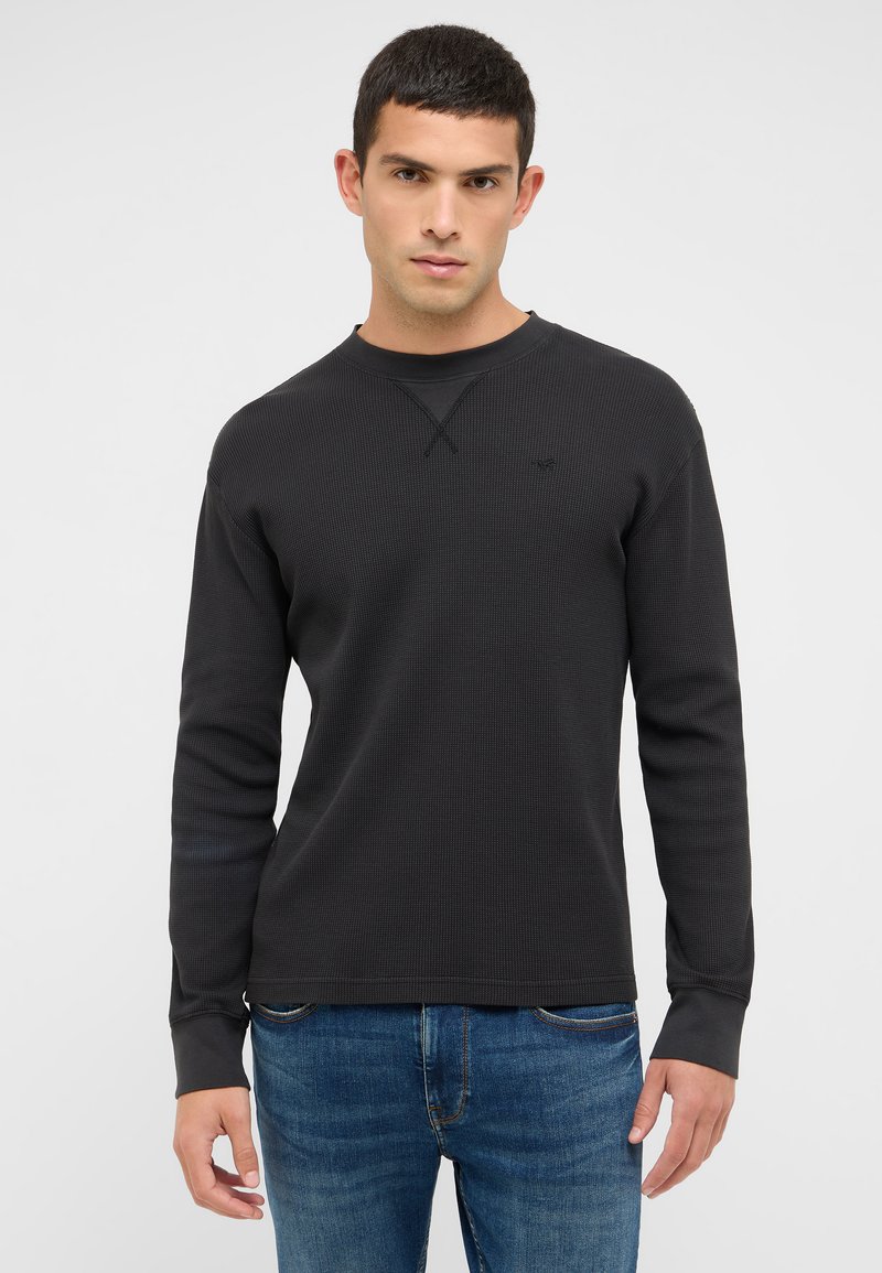 Fekete hosszú ujjú, gofré-kötött póló kerek nyakkal, finom logóval és texturált mintával. Kék farmernadrággal viselve.