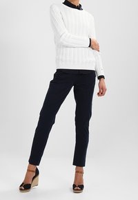 Bílý svetr s pleteným vzorem, námořnické kalhoty a hnědé sandály na podpatku s černými akcenty. Oblečení se vyznačuje texturovanou tkaninou a přiléhavým střihem.