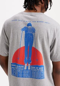 Camiseta gris con un gráfico azul de un esquiador sobre un círculo rojo. El texto incluye "LEVI'S SKI RODEO" y detalles del evento, impresos en colores contrastantes.
