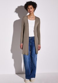 Cardigan lungo aperto in tessuto verde oliva, indossato sopra una maglietta bianca, abbinato a jeans a gamba larga blu e sneakers bianche. Texture liscia, design minimal.