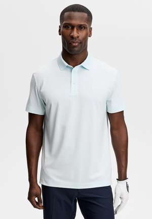 Mann trägt ein hellblaues kurzärmeliges Poloshirt, dunkle Hose und einen weißen Golfhandschuh an der linken Hand, steht vor einem schlichten Hintergrund.