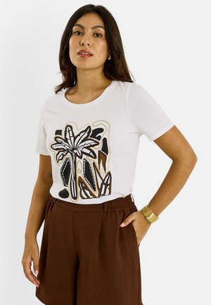 Mujer con camiseta blanca con diseño abstracto de hojas negras y marrones, pantalones cortos marrones de cintura alta, pulsera de oro, de pie con la mano en el bolsillo.