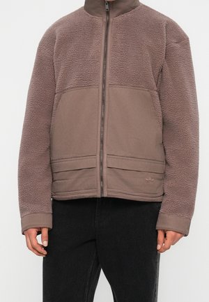 Personne portant une veste polaire marron zippée avec des panneaux en tissu lisse et une polaire texturée sur les manches et le haut de la poitrine.