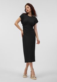 Černé midi šaty s krátkými rukávy, bočním nařasením a přiléhavým střihem. Modelka má na sobě zlaté platformové podpatky. Hladká textilie.
