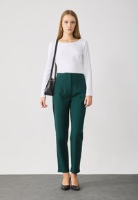 Witte blouse met lange mouwen, groene op maat gemaakte hoog onderste broek, zwarte hakken en een kleine zwarte tas. Glad materiaal met geplooide details.
