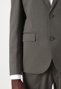 Veste de costume grise sur mesure avec un revers cranté, fermeture à deux boutons et poche latérale. Texture lisse avec des détails de couture visibles.
