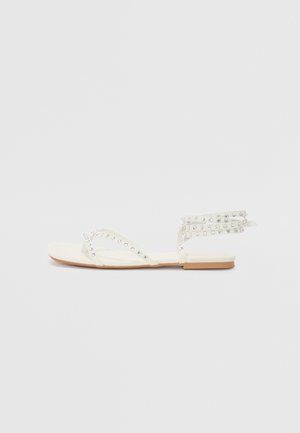 Sandales plates blanches avec des accents décoratifs en strass, présentant des brides croisées et une sangle à la cheville. Texture lisse avec une semelle marron clair.