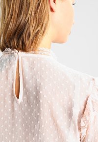 Jasno różowa bluzka z strukturalnym wzorem w kropki. Posiada koronkowe wykończenie dekoltu oraz otwarcie w kształcie łezki z tyłu. Rękawy z falbanami.